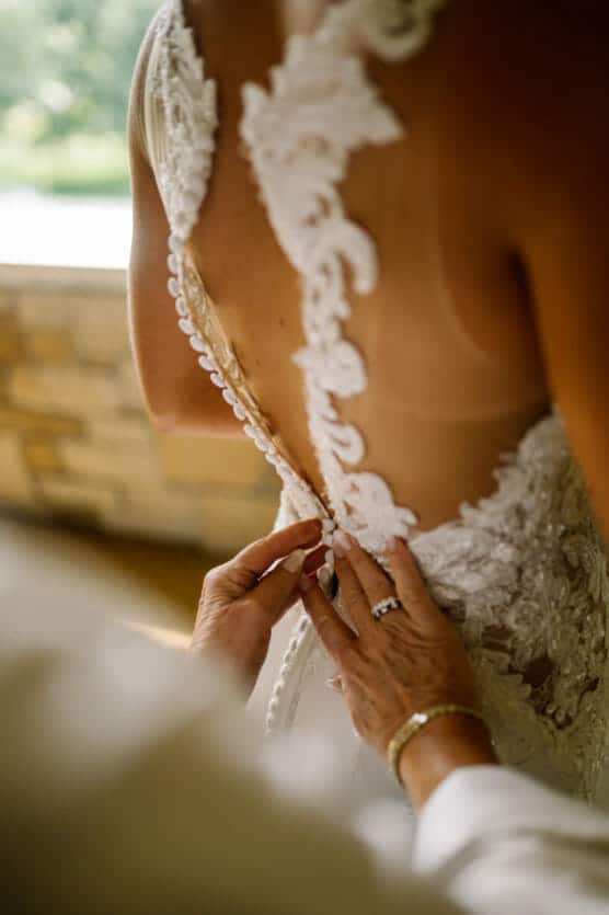 Close-up of someone buttoning the back of a lace wedding dress on a bride, focusing on the intricate details of the gown and the gentle hands fastening the buttons. by Justin Salem Meyer