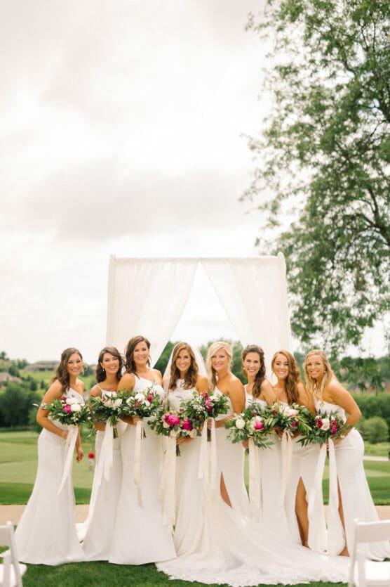 Eight women in white dresses stand outdoors, smiling and holding floral bouquets in front of a sheer white wedding arch, with green grass and trees in the background. by Justin Salem Meyer