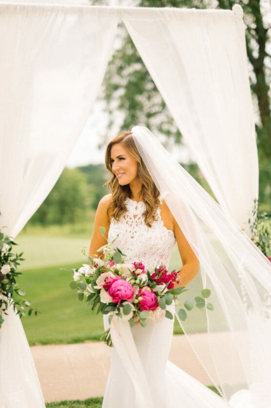 A bride in a white lace wedding dress stands outdoors under sheer white drapes, holding a bouquet of vibrant pink and white flowers, with greenery in the background. She smiles and looks to the side. by Justin Salem Meyer