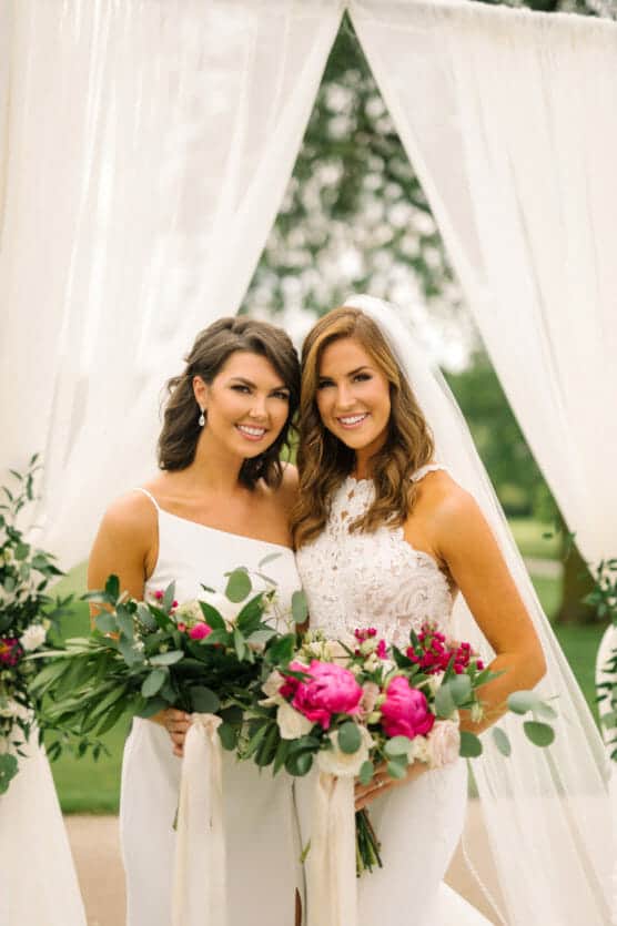 Two women in white dresses stand side by side, smiling and holding bouquets with greenery and pink flowers. A sheer white curtain and greenery create a soft, elegant backdrop at an outdoor wedding. by Justin Salem Meyer