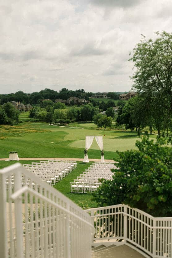 A wedding ceremony setup on a golf course with rows of white chairs facing a white draped archway, surrounded by lush green grass and trees under a cloudy sky. by Justin Salem Meyer