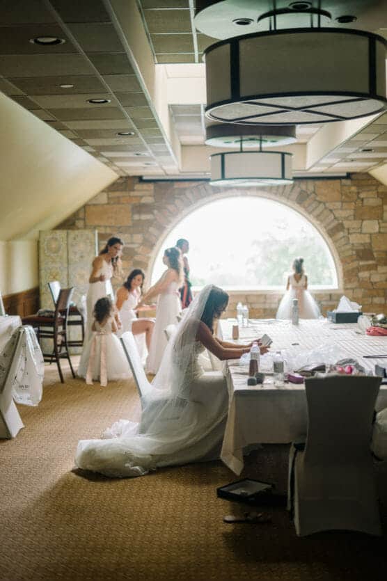 A bride in a white dress sits at a table fixing her makeup, while bridesmaids and flower girls in light dresses get ready in a softly lit room with a large arched window and stone wall in the background. by Justin Salem Meyer