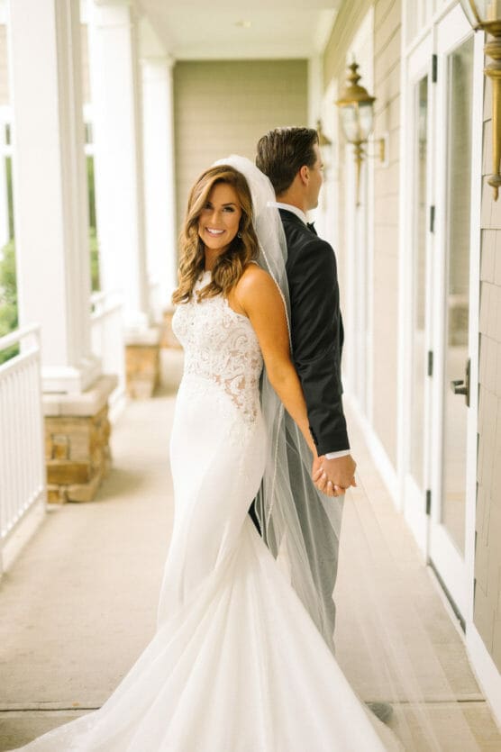 A bride in a white gown and veil smiles at the camera while standing back-to-back and holding hands with a groom in a black suit on a bright, outdoor porch. by Justin Salem Meyer