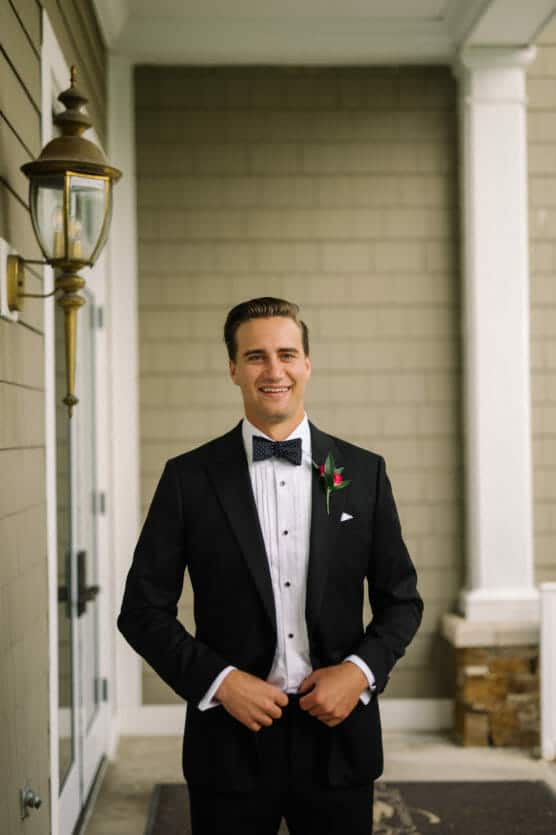 A man in a black tuxedo with a bow tie and a boutonniere stands smiling in front of a tan building with white trim and a glass door, posing for a formal photo. by Justin Salem Meyer