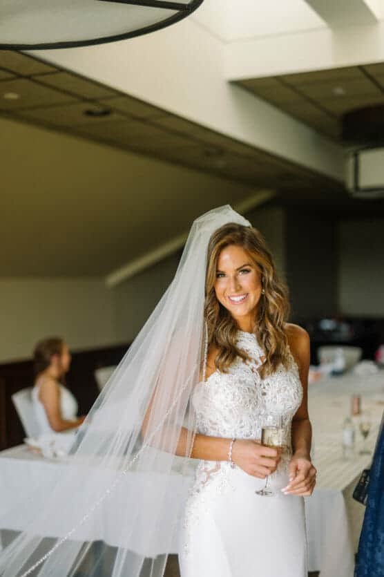 A smiling bride in a white lace wedding dress and long veil holds a glass of champagne indoors, with another woman seated at a table in the background. by Justin Salem Meyer