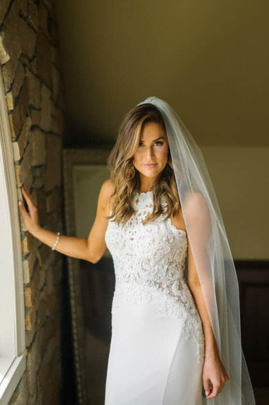A bride in a white lace wedding dress and long veil stands by a stone wall, looking at the camera with soft lighting highlighting her face and hair. by Justin Salem Meyer