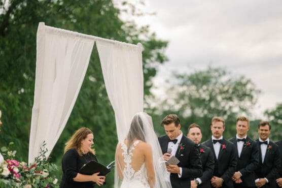 A bride and groom stand under a white draped arch outdoors during their wedding ceremony, with the officiant reading from a book and groomsmen in black suits standing in the background. Trees and greenery surround the scene. by Justin Salem Meyer
