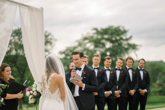 A groom in a black tuxedo reads vows to his bride, who wears a white dress with a lace back, during an outdoor wedding ceremony. Groomsmen stand in a row behind him, and a smiling officiant holds a book nearby. by Justin Salem Meyer