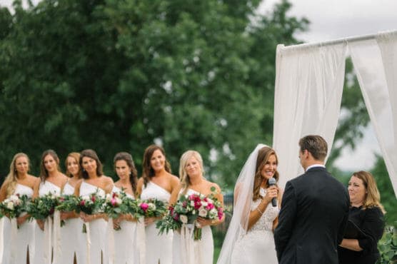 A bride in a white dress and veil smiles as she speaks into a microphone facing the groom during an outdoor wedding, with bridesmaids in matching dresses holding bouquets standing nearby. by Justin Salem Meyer