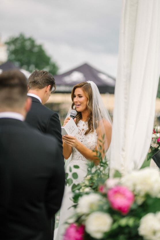 A bride in a white dress and veil reads vows from a booklet into a microphone during an outdoor wedding ceremony, facing the groom. Pink and white flowers are in the foreground. by Justin Salem Meyer