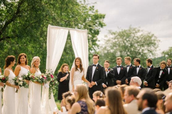 A bride and groom stand under a white wedding arch outdoors, surrounded by bridesmaids in white dresses holding bouquets and groomsmen in black suits with bow ties, as guests watch the ceremony. by Justin Salem Meyer