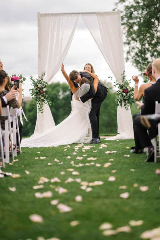 A newlywed couple shares a joyful kiss under a white arch outdoors, surrounded by guests sitting on either side of a petal-strewn aisle. The bride raises her bouquet in celebration. by Justin Salem Meyer