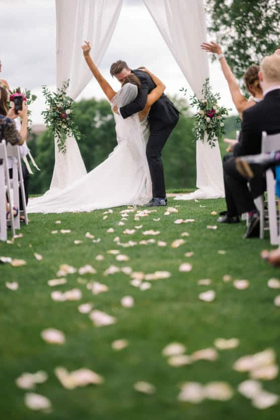 A bride and groom kiss passionately at an outdoor wedding ceremony under a white arch, surrounded by guests seated on either side and flower petals scattered on the grass aisle. by Justin Salem Meyer