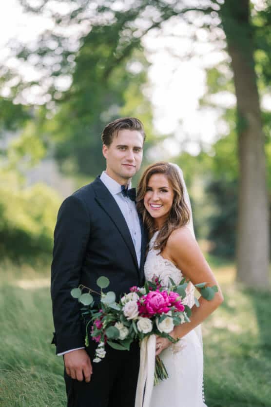 A bride and groom stand close together outdoors, smiling at the camera. The groom wears a black tuxedo and bow tie; the bride wears a white dress and holds a bouquet of pink and white flowers. Green trees blur in the background. by Justin Salem Meyer