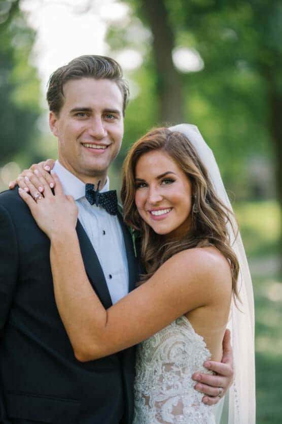 A bride in a lace dress and veil embraces a groom in a tuxedo outdoors, both smiling at the camera with greenery in the blurred background. by Justin Salem Meyer