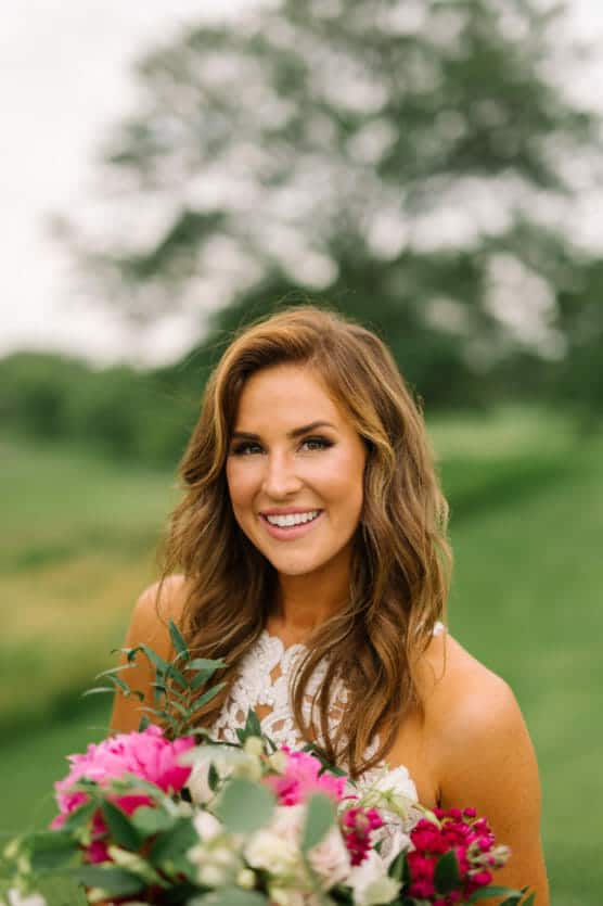 A woman with wavy brown hair smiles outdoors, holding a colorful bouquet of flowers. She wears a white lace dress, and green trees and grass are blurred in the background. by Justin Salem Meyer