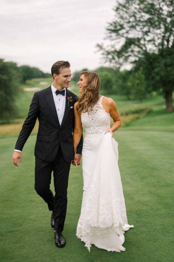A bride in a white gown and a groom in a black tuxedo walk hand in hand on green grass, smiling at each other, with trees and a cloudy sky in the background. by Justin Salem Meyer