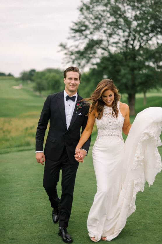 A bride in a white dress and a groom in a black tuxedo walk hand-in-hand on a grassy field, smiling and looking happy on their wedding day, with trees in the background. by Justin Salem Meyer