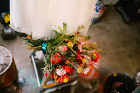 A colorful bouquet of flowers with yellow, red, and pink blooms rests on a metallic chair beneath a white draped fabric, set on a concrete floor with various objects in the background. by Justin Salem Meyer