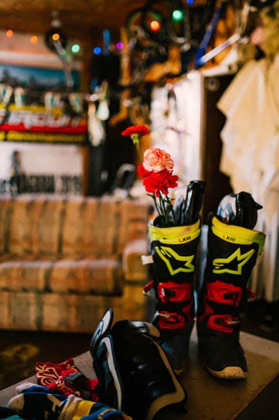 A pair of motocross boots with red and pink carnations inside are on a table in a cozy, colorful room. A sofa, bikes, and string lights are visible in the warmly lit background. by Justin Salem Meyer