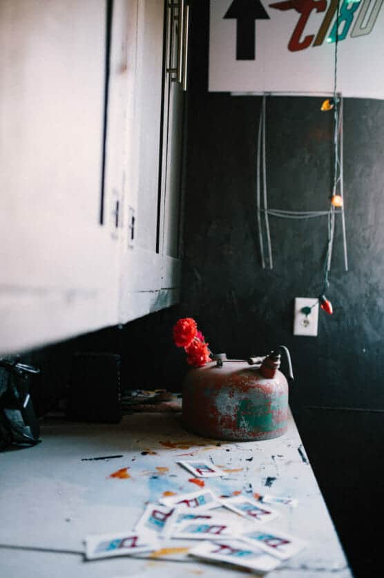 A rusty red kettle with a red artificial flower sits on a cluttered countertop, surrounded by scattered cards. In the background, string lights hang near a wall outlet and a partially visible poster. by Justin Salem Meyer