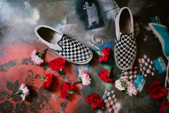 A pair of black-and-white checkered slip-on shoes on a colorful, spray-painted surface, surrounded by scattered red and pink carnations. by Justin Salem Meyer