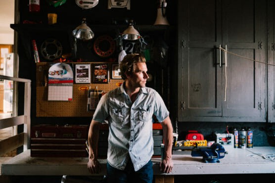 A person in a short-sleeve button-up shirt stands in a workshop, leaning against a workbench. Shelves, tools, calendars, and various workshop items are visible in the background. Light shines in from the left. by Justin Salem Meyer