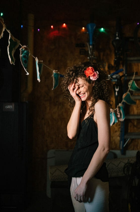 A woman with curly hair and a flower in her hair smiles and covers her face with one hand, standing indoors under colorful string lights and festive decorations. by Justin Salem Meyer