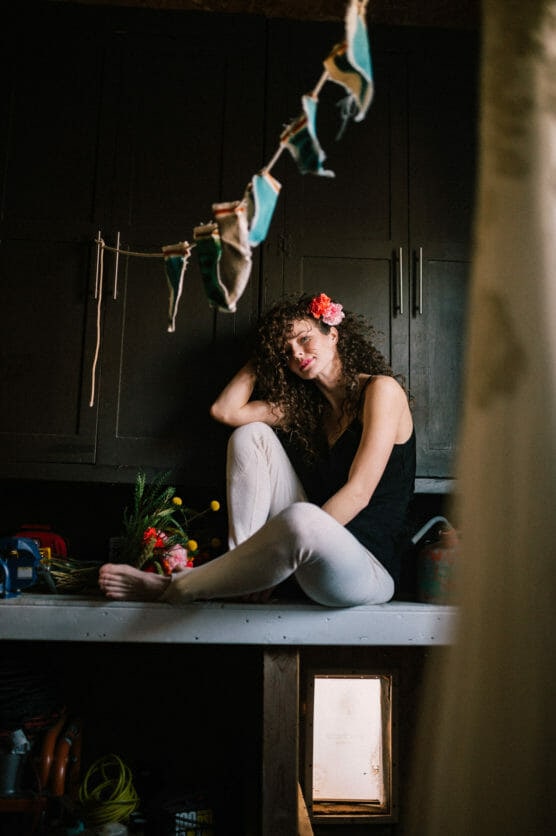 A woman with curly hair and a flower in her hair sits on a countertop, barefoot, with her knees up and head resting on one hand. Colorful bunting hangs in the foreground and a small open hatch is visible below her. by Justin Salem Meyer