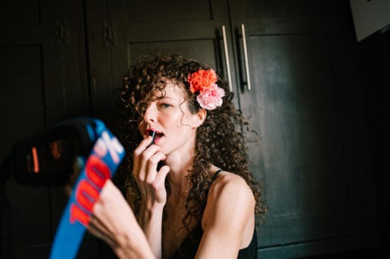 A woman with curly hair and pink and red flowers in her hair applies lipstick while looking into a mirror. She is sitting in front of dark-colored cabinets. by Justin Salem Meyer