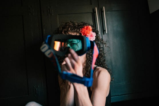 A person with curly hair and a red flower accessory holds a blue and orange pair of goggles in front of their face, partially obscuring it, with dark cabinets in the background. by Justin Salem Meyer