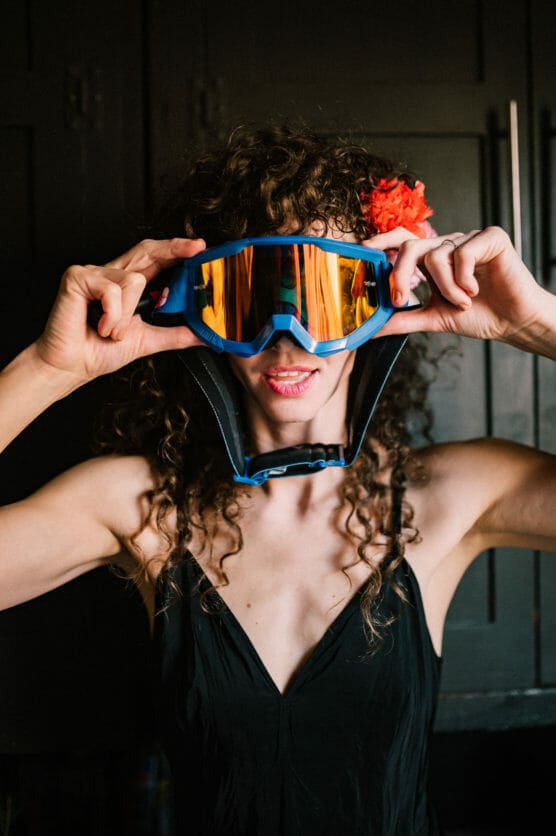 A person with curly hair and a red flower tucked behind their ear adjusts blue and orange ski goggles while wearing a black sleeveless top, standing indoors against dark cabinets. by Justin Salem Meyer