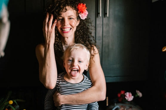 A woman with curly hair and a flower in her hair smiles while hugging a young child with blonde hair, who is sticking out their tongue. Both are wearing striped shirts and are standing in a dark room with flowers in the background. by Justin Salem Meyer