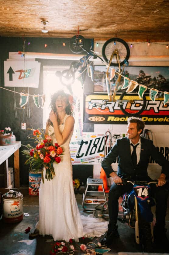 A joyful bride in a white dress and holding a colorful bouquet stands next to a groom in a suit sitting on a small dirt bike in a garage decorated with banners, bikes, and racing posters. by Justin Salem Meyer