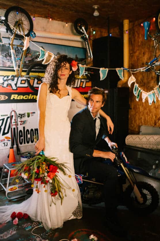 A bride in a white dress stands holding a bouquet, while a groom in a suit sits on a small motorcycle. They are in a decorated garage with bikes, banners, and string lights. The mood is playful and unconventional. by Justin Salem Meyer