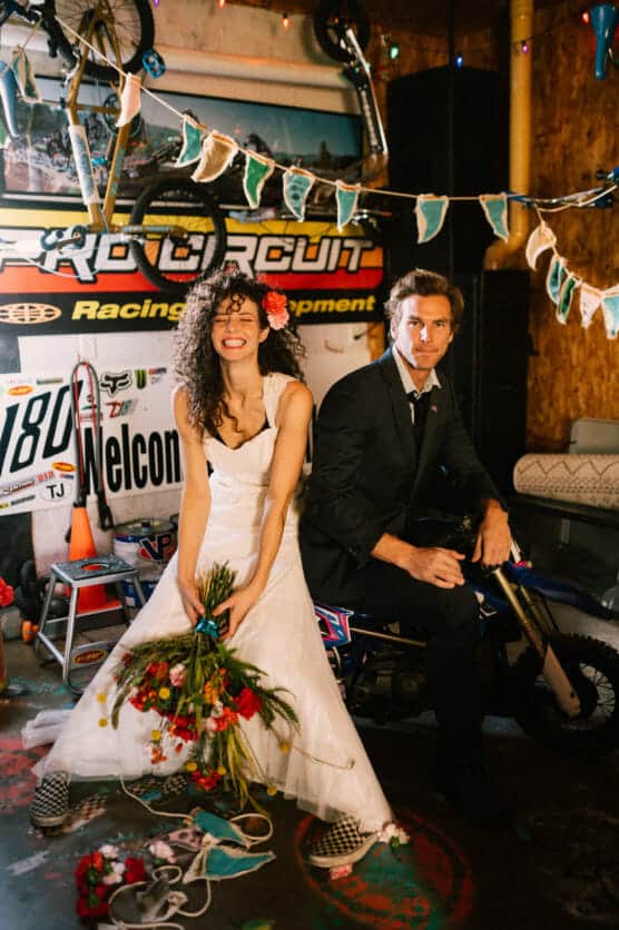 A bride in a white dress and flower crown sits playfully on a mini dirt bike holding a bouquet, while the groom in a suit sits behind her. They are in a garage decorated with racing gear and string lights. by Justin Salem Meyer