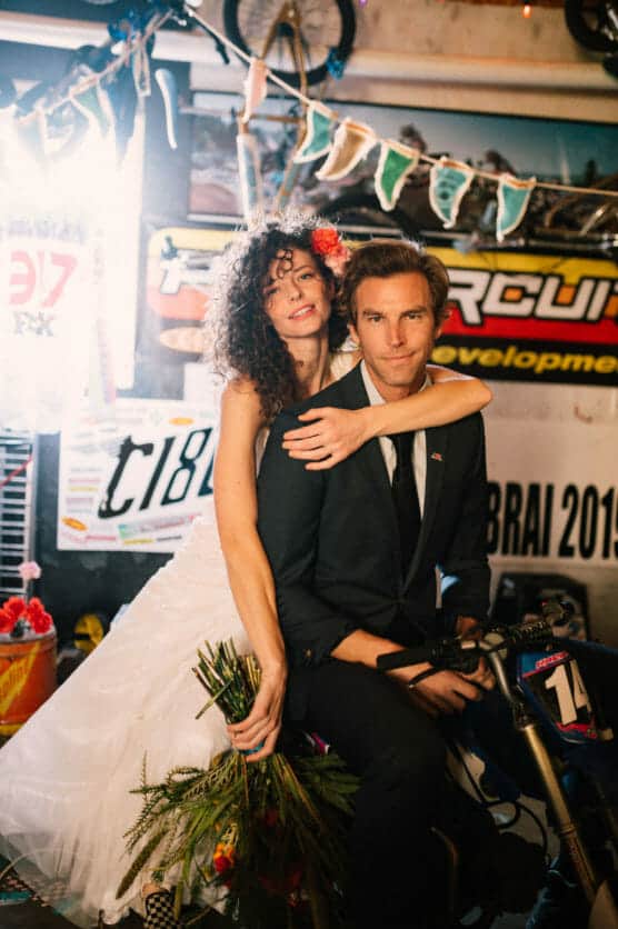 A bride in a white dress and a groom in a suit sit together on a small motorbike indoors. The bride holds a bouquet and smiles, while the groom looks at the camera. The background has racing posters and decorations. by Justin Salem Meyer