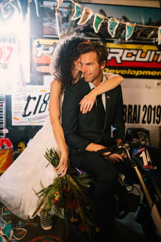 A bride in a white dress embraces and kisses a groom in a black suit from behind as they sit together on a small motorcycle, surrounded by colorful decorations and banners. by Justin Salem Meyer