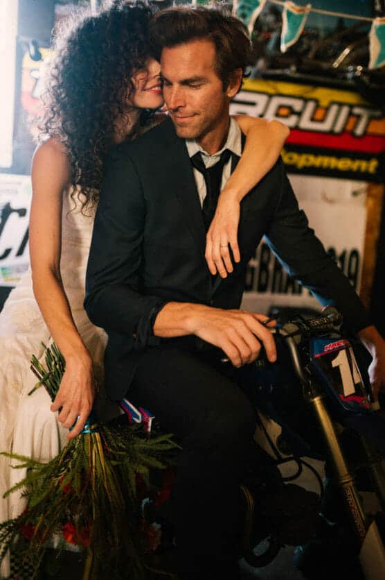 A woman in a white dress sits on a small motorcycle behind a man in a black suit, both smiling closely. The woman holds a bouquet, and racing signs are visible in the background. by Justin Salem Meyer