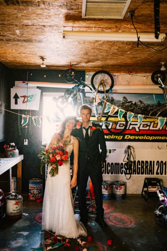 A bride in a white gown holding a bouquet leans on a groom in a suit. They stand in a garage decorated with bike parts, tools, banners, and string lights under a Racing Development sign. by Justin Salem Meyer