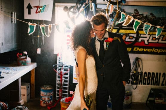 A bride in a white dress and a groom in a suit smile and lean close together in a decorated garage with banners, racing posters, and bicycles in the background. by Justin Salem Meyer