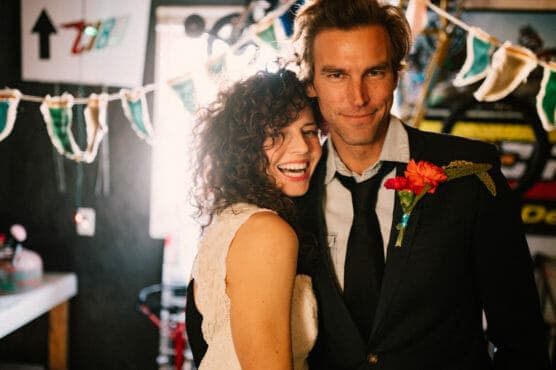 A smiling couple stands close together at a festive indoor party. The woman has curly hair and wears a white dress; the man wears a suit with a red flower boutonniere. Colorful pennant banners hang in the background. by Justin Salem Meyer