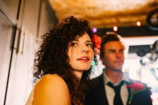 A woman with curly hair and a red flower tucked behind her ear stands next to a man in a suit and tie, both indoors with warm lighting and blurred festive lights in the background. by Justin Salem Meyer