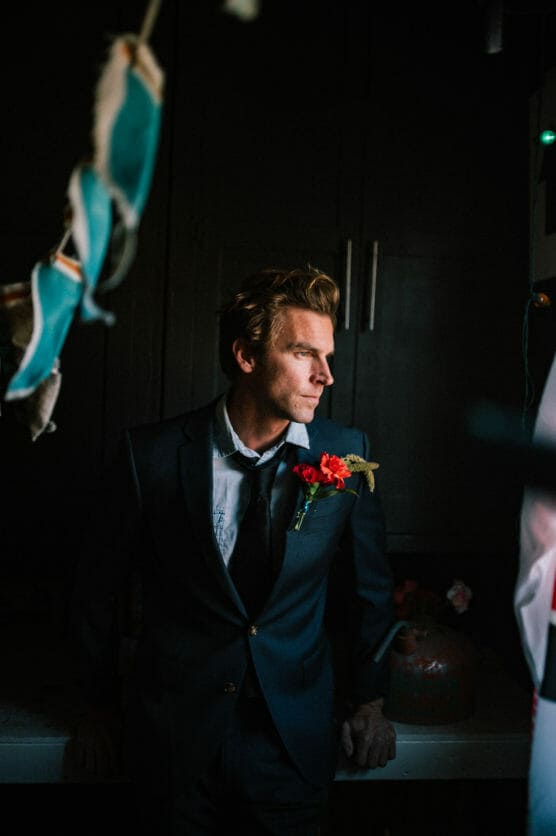 A man in a dark suit with a red flower boutonniere stands in a dimly lit room, looking to his left. Light shines on his face, highlighting his thoughtful expression, while the background remains shadowy. by Justin Salem Meyer