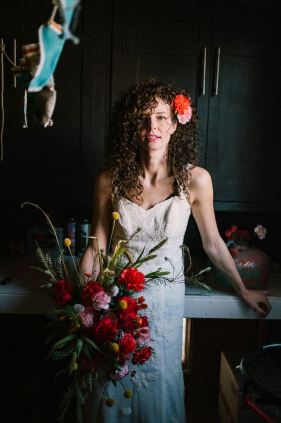 A woman in a white lace dress with curly hair and a red flower hairpiece stands indoors, holding a colorful bouquet of red and pink flowers, with dark cabinets and shelves in the background. by Justin Salem Meyer