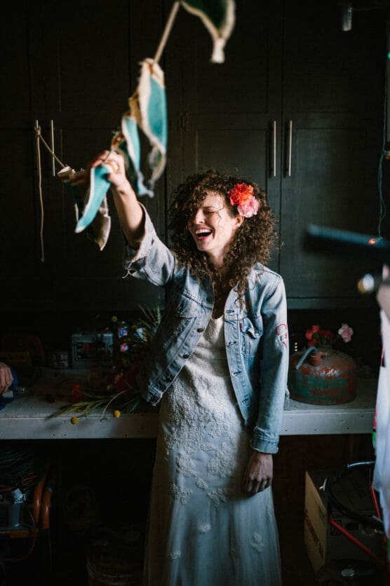 A woman with curly hair, wearing a denim jacket over a white dress and a red flower in her hair, smiles joyfully and holds up a decorative garland in a dimly lit room with cabinets and crafts on the counter. by Justin Salem Meyer