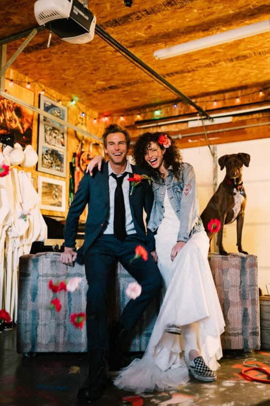 A joyful bride and groom sit together in a decorated garage, laughing as flower petals fall around them. The bride wears a white dress, jean jacket, and checkered shoes, while a dog sits nearby on a bench. by Justin Salem Meyer