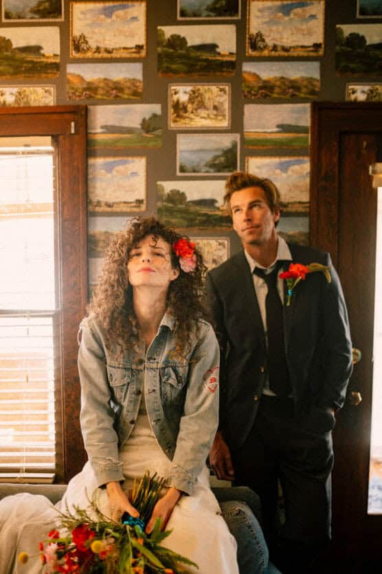 A woman in a denim jacket and white dress sits holding flowers, with a red flower in her curly hair. A man in a suit with a loosened tie stands beside her. They are indoors, with landscape paintings on the wall behind them. by Justin Salem Meyer