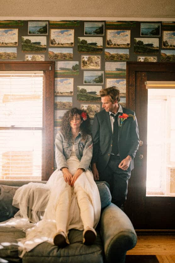 A woman in a wedding dress and denim jacket sits on a couch, smiling, next to a man in a suit. They both wear red flower boutonnieres. The room has vintage-style paintings on the wall behind them. by Justin Salem Meyer
