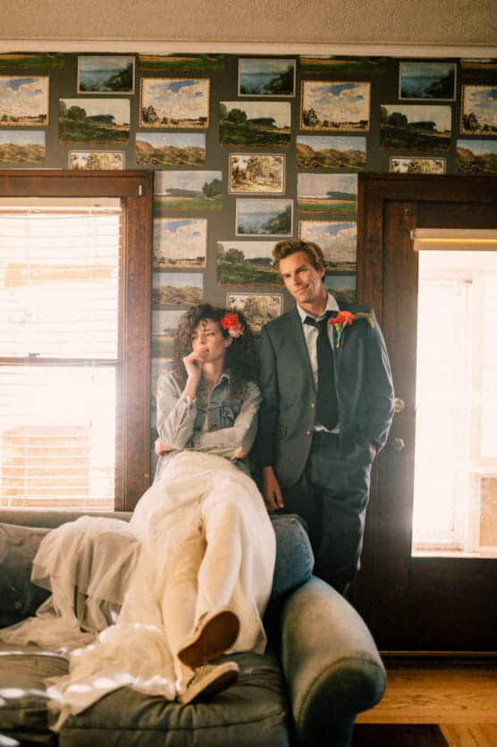 A bride in a white dress and denim jacket lounges on a sofa, looking thoughtful, while a groom in a suit stands beside her, both with flower boutonnieres, in a cozy room with landscape paintings on the wall. by Justin Salem Meyer